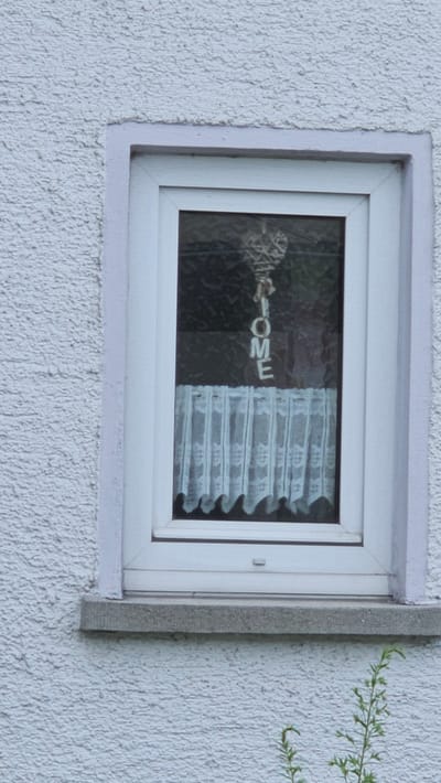A white, textured exterior wall of a building in Vöhringen, Germany, surrounds a white-framed window. The windowpane is dark, obscuring the interior, but a white string of letters spelling "HOME" hangs from the top of the window. Below the word, a white lace curtain with a scalloped hem is visible. A grey concrete windowsill sits beneath the window. In the foreground, a small green plant with thin stems and leaves grows near the base of the wall.
