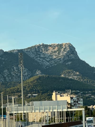 The image captures a daytime scene in the city of Toulon, France, dominated by the prominent Mont Faron mountain. The mountain's rugged, rocky slopes are partially covered with green vegetation and trees. At the summit of Mont Faron, several structures, possibly observation points or antennas, are visible against the clear blue sky.

In the foreground, a tall, slender lighting tower stands on the left side of the image. Below the mountain, a suburban or urban landscape unfolds, featuring several multi-story buildings. One building, light beige in color, has a distinctive modern architectural style with visible antennas on its roof. Adjacent to it, a longer, more contemporary building with numerous windows is also present. A white truck is visible on the right side of the image, suggesting it is on a road or highway. Barriers and streetlights are also part of the foreground infrastructure, indicating a road or public area. The overall environment appears to be a mix of natural landscape and urban development.