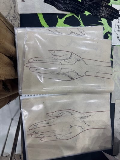 The image displays two sheets of fibrous, light brown paper, each encased in a clear plastic sleeve, exhibiting linear drawings of stylized human left hands. The sheets are positioned vertically, with the top sheet partially overlapping the one below it. The lower sheet's plastic sleeve reveals three circular perforations along its left edge, indicating it is part of a binder or display album. Each drawing features a hand outline rendered in a thin reddish-brown line, with intricate black curvilinear patterns and lines detailing the palm and wrist areas. The scene is located in Ban Rong Ruea Kham, Thailand. To the immediate left of the displayed pages, a section of brown, textured fabric is visible. Above the drawings, a black textile with a bright neon green abstract or organic pattern is present. Overlying this patterned fabric, a black, hand-shaped object adorned with silvery-white intricate wavy designs is visible, alongside a partial white label featuring black stylized script. The environment appears to be indoors, under ambient lighting conditions that create some glare on the plastic sleeves.