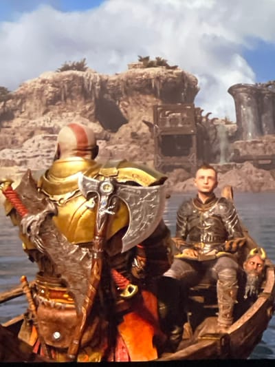 The image shows Kratos and Atreus in a boat, likely from the video game "God of War." Kratos is in the foreground, facing left, with his Leviathan Axe prominently displayed. He wears golden armor. Atreus stands behind him, gazing directly at the viewer, wearing similar armor. The background reveals a rocky shoreline and a partially visible building or structure. The sky is cloudy.