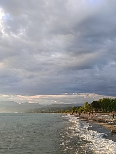 A wide shot of a rocky beach in Gulrypsh, Abkhazia, features a calm sea with gentle waves lapping at the shore under a cloudy sky. In the distance, rolling mountains form a hazy backdrop. Several people are scattered along the beach, some walking and others standing, while a few are sitting. The beach is covered with pebbles and driftwood. The overall scene depicts a serene coastal landscape with human presence.