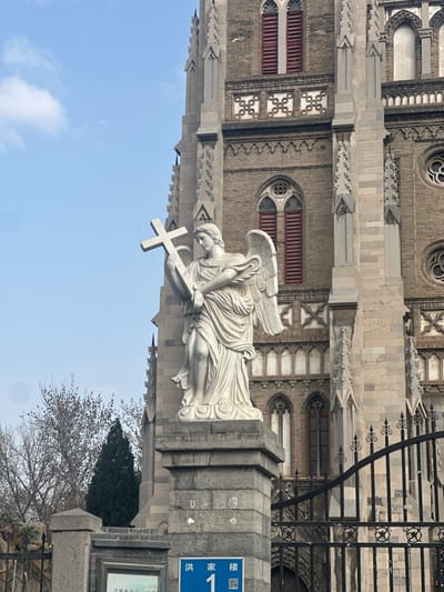 This image captures the exterior of Hongjialou Cathedral (Cathedral of the Sacred Heart of Jesus), located in Licheng District, Jinan, China. A blue street sign in the lower left corner clearly displays "洪家楼 1" (Hongjialou 1), confirming the address.

In the foreground, a white marble or stone statue of an angel stands on a multi-tiered grey block pedestal. The angel is depicted with large wings, draped classical garments, and holds a plain Latin cross in its right arm, resting it against its upper torso. Its head is slightly bowed and turned to the left. The pedestal itself is part of a larger grey stone block pillar or wall segment.

To the right of the statue, a black wrought-iron fence or gate with decorative scrollwork partially frames the scene and obscures the lower section of the cathedral. The background is dominated by the large, multi-story Gothic-revival style cathedral, constructed from weathered grey and brown brick. Visible architectural features include multiple pointed arch windows fitted with red louvered blinds, intricate stone tracery, decorative patterned brickwork, and buttresses supporting the structure. Elements of tall spires or pinnacles are visible along the cathedral's roofline.

On the left side of the frame, behind the statue, bare deciduous trees and a dark green coniferous tree are visible, suggesting a cooler season. The upper-left portion of the sky is clear light blue with a few sparse white clouds. No people are visible in the scene.