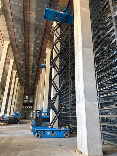 The image depicts the interior of a large building under construction. Overhead, a network of metal support beams forms the ceiling structure, with concrete beams running perpendicular to the metal grid. Two blue scissor lifts, possibly Genie brand, are positioned between the concrete beams, extending towards the ceiling. Below, long rows of gray rectangular objects line the space between concrete beams, possibly air ducts or similar mechanical systems. Red pipes run along the lengths of these rows. The overall scene suggests a large industrial or commercial building in progress, with the scissor lifts likely used for installing the ceiling or upper-level infrastructure. While specific location cannot be determined, it is NOT in the city of Caesarea, Israel.