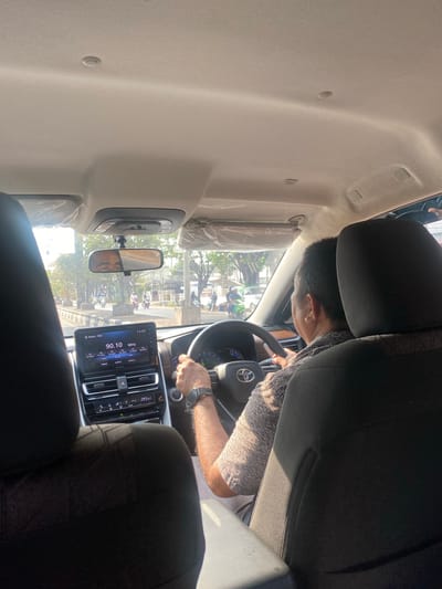 The image shows the interior of a car looking forward from the back seat. A man is driving, his hands on the steering wheel. The car's dashboard is visible, featuring a touchscreen infotainment system displaying the radio station "Radio - FM1" and the time "14:52." Sunlight streams in from the windows, illuminating the interior. Outside, a street scene unfolds with trees, other vehicles, and a building. The overall impression is of a daytime drive on a paved road, possibly in a suburban or urban environment. The specific location of Mandar Sari, Indonesia, is not explicitly indicated within the image itself.