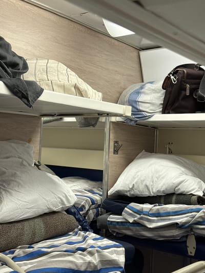 The image depicts a cramped interior, likely a train compartment, with bunk beds. On the upper bunk, a dark grey garment is draped over the edge, and a striped pillow is visible. Adjacent to it, a brown leather bag rests on a shelf. The lower bunk is more cluttered. A large white pillow sits atop a rolled-up blanket with a textured, speckled pattern. Below this, striped bedding in blue, grey, and white covers the mattress. The walls are paneled with a light wood-grain laminate. Metal fixtures, possibly coat hooks, are attached to the partition between the bunks. The lighting is dim, suggesting nighttime or an interior space. The overall impression is one of a confined, utilitarian sleeping area. The location is specified as Байлянгар, Russia.