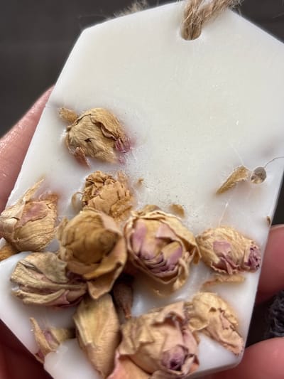 The image shows a close-up view of a white wax sachet or melt embedded with several dried rosebuds. The wax appears to be translucent in places, allowing the color of the rose petals to show through. A rough jute string is threaded through a hole at the top of the sachet. The sachet is held by a person's hand, with only the fingers and thumb visible. The background is dark and out of focus. The scene does not contain any elements that can be used to determine its location in Novaya Usman, Russia.