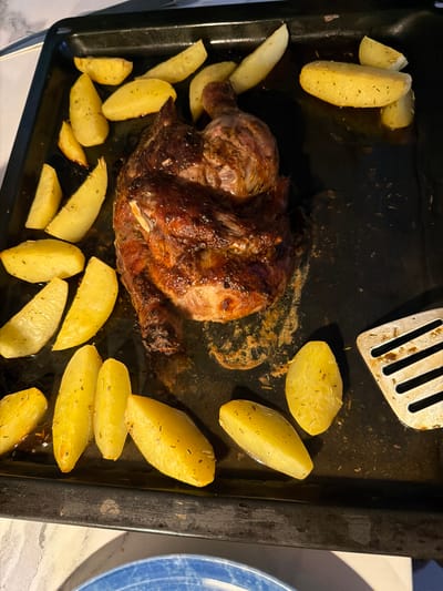 In Novoalexandrovka, Russia, a roasted chicken and potato dish is presented on a dark baking sheet. The chicken, browned and glistening with juices, is positioned centrally. Scattered around the chicken are numerous golden-brown potato wedges, seasoned with herbs. A silver spatula rests on the right side of the baking sheet, near a cluster of potatoes. The baking sheet itself appears used, with some food residue evident. The overall presentation suggests a home-cooked meal.