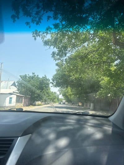 The image is taken from inside a vehicle, looking through the windshield. The foreground shows the dark interior of the car's dashboard. The middle ground depicts a street lined with trees, a few buildings, and a utility pole. On the left side of the road, there is a small white building with a blue-trimmed window and a wooden gate. Further down the street, several cars and some pedestrians can be seen. The background shows a clear blue sky with some tree branches and leaves visible at the top, suggesting a sunny day. The scene appears to be a residential area in a town or village, possibly in a rural part of Kyrgyzstan, given the style of the buildings and vegetation.