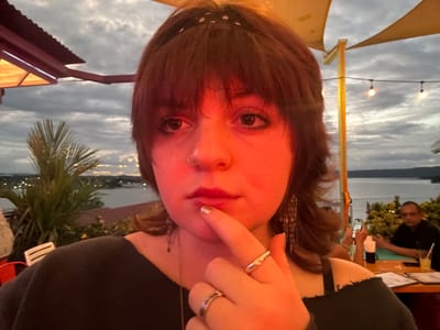 An upside-down selfie of a fair-skinned person with brown hair and bangs. They have brown eyes and are wearing jewelry including rings, earrings, and a nose ring. Their finger is near their nose. In the background, a body of water and a skyline are visible, along with outdoor restaurant decor, a palm tree, and other people.
