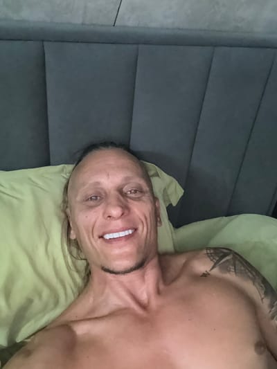 A shirtless man with long, dark hair pulled back lies in bed, propped up on a light green pillow against a grey headboard. He smiles at the camera, revealing white teeth. A tattoo is visible on his upper right arm. The background suggests an indoor setting, possibly a bedroom in Vol'noe, Russia.