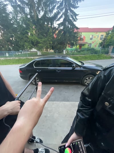 In Tovstoluh, Ukraine, a black sedan is parked on a paved area next to a grassy strip and a fence. Beyond the fence, a two-story building with a red roof and a green facade featuring abstract patterns is visible. Several tall evergreen trees are positioned in front of the building and fence. In the foreground, three people are partially visible. One person on the left is holding a metallic object, and another person in the center is holding up two fingers in a peace sign. The person on the right is holding a smartphone. The ground is paved with concrete.