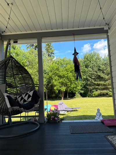 The image depicts a view from a covered porch looking out onto a grassy backyard. In the foreground, a dark grey woven hanging chair with black and white patterned cushions and a throw blanket is visible on the left. To the right, on the porch floor, are two dark grey mats and a pink cushion. A potted plant with pink flowers sits near the edge of the porch.
Suspended from the porch ceiling by a red string is a decorative witch with a black pointed hat, a black dress, and multicolored streamers. In the background, a green lawn is dotted with trees and bushes. Beyond the lawn, a bright blue sky with scattered white clouds is visible. On the lawn, a grey lounge chair and a colorful inflatable object are present. A small statue of a seated Buddha is located on the porch to the right, near the edge of the lawn. The scene appears to be a residential setting, possibly a backyard in Алешкино Дп, Russia, during daylight hours.