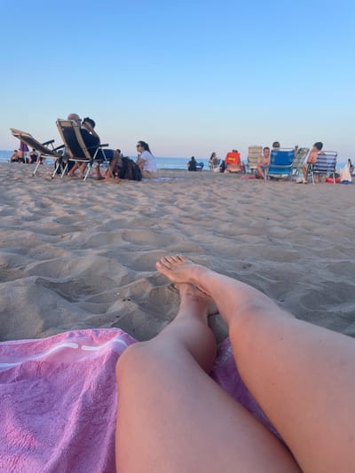 This image depicts a beach scene at Canet d'en Berenguer, Spain, during the late afternoon or early evening, indicated by the warm lighting and clear blue sky. The foreground features a person's legs and bare feet resting on a pink towel laid out on the sandy beach. The sand is textured with ripples, suggesting a gentle breeze. In the mid-ground, several beach chairs are occupied by people. Some are sitting and interacting, while others appear to be relaxing. The background shows the calm sea meeting the horizon.