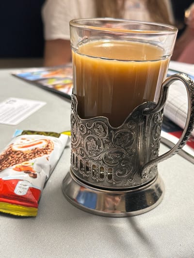 An indoor scene in Kostino, Russia, displays a glass cup containing a light brown beverage, likely coffee or tea with milk, seated within an ornate, silver-colored metal podstakannik featuring embossed floral and leaf motifs. To the left, a partially open Kinder-brand snack wrapper, predominantly white and red, displays an image of a chocolate bar with nuts and caramel, along with Russian text. All items rest on a light grey, subtly textured fabric surface. The background is out of focus, revealing blurred figures of individuals and indistinct papers or booklets on an adjacent surface. No direct human action is depicted within the frame.