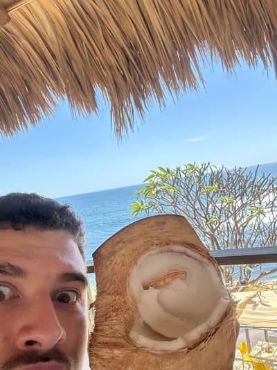 From a tilted perspective, a man’s face peers into the camera, partially obscured by a halved coconut held aloft. The coconut reveals white flesh. Behind him, the backdrop showcases a serene blue sea and sky, partially framed by a tree with sparse green leaves. A thatched roof extends from the left edge of the frame, adding a tropical ambiance to the scene.
