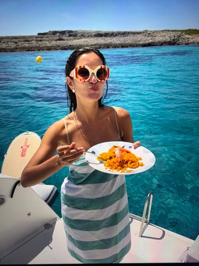 The image is oriented sideways, rotated 90 degrees counter-clockwise. It depicts a woman on a boat enjoying a meal amidst clear, calm turquoise waters. The woman, with dark hair and distinctive red and white striped sunglasses, is holding a white plate filled with an orange-yellow dish, likely paella, garnished with a large shrimp, and a fork. A green and white striped towel is draped around her, suggesting a relaxed setting. She is looking towards the camera with a slightly pouting, playful expression.

In the background, a rugged, rocky coastline meets a bright blue sky, enhancing the serene marine environment. The bright daylight indicates it is midday. A small yellow object floats in the distant water. Partially visible near the boat is a white paddleboard or surfboard with reversed red text that reads 'ełmo O.' and 'eimio' (when viewed in the current orientation). The scene, characteristic of a warm and sunny climate, is set with the provided context of Ros Na Rí, Ireland.