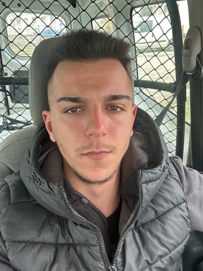 This image depicts a male individual within a vehicle in Srednia, Bulgaria. The subject, appearing to be a young adult male in his early to mid-twenties, is shown from the chest up, directly facing the camera. He possesses short, dark hair, brown eyes, a straight nose, and faint stubble on his upper lip and chin. He is dressed in a dark grey quilted puffer vest worn over a darker, possibly long-sleeved, garment. The individual is seated in a light grey upholstered vehicle seat. Behind him, a metal mesh security partition is visible, separating the front of the vehicle from a rear compartment. Through the vehicle's windows and the mesh, an exterior scene is discernible, featuring blurred residential or commercial buildings, a white vehicle, and industrial machinery, possibly an excavator with blue elements, under an overcast sky.