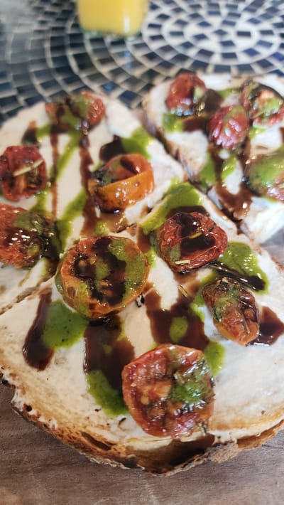 The image depicts a plate of bruschetta, featuring toasted bread topped with a white spread (likely ricotta or goat cheese), roasted tomatoes, green pesto, and balsamic glaze. The food is arranged on a mosaic patterned surface, with a wooden countertop partially visible. The scene is indoors in the city of Jackson, United States of America.