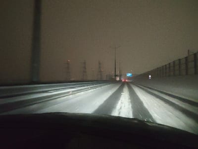 The image captures a nighttime, forward-facing view from inside a moving vehicle on a multi-lane highway in Belyaninovo, Russia. The lower portion of the vehicle's dashboard is visible. The road surface is partially covered in snow and ice, with the vehicle's headlights illuminating distinct, wet, clear tire tracks in the immediate foreground and mid-distance. The sky is dark, overcast, and hazy, suggesting snowfall or fog, limiting long-distance visibility.

To the left, a prominent dark vertical structure, possibly a pole or support, stands in the foreground. Further in the background on the left, several large electrical transmission towers, each with a red warning light, are discernible against the dim sky. To the right, a tall, continuous noise barrier or retaining wall, light in color, runs parallel to the highway. A circular blue road sign with a white border is affixed to this wall. Ahead, various streetlights illuminate sections of the road, and an overhead blue road sign with white text is visible. The red tail lights of other vehicles are present in the distance, indicating traffic ahead on the highway. No people are visible.
