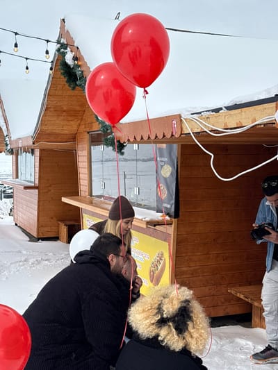 This image captures a winter scene in Yeghipatrush, Armenia, featuring a group of individuals gathered around wooden kiosks on snow-covered ground. The sky is overcast and white.
In the foreground, two large, glossy red balloons float upwards, tethered by red ribbons, one ribbon extending towards a person in the foreground. A white balloon is partially visible behind one of the individuals. The ground is uniformly covered in snow.
Two wooden kiosks with peaked roofs, also covered in snow, are visible. Green garlands are draped beneath the eaves of the kiosks. String lights are strung between the kiosks. The kiosk on the right features a serving counter and a yellow sign displaying "THIN hot dog" and Armenian text, along with an image of a hot dog. Another sign above it displays "MASUN" and "URAN" with graphic elements. White electrical wiring is visible along the roofline of the rightmost kiosk.
Four individuals are present. In the center-left, a male with short dark hair and glasses wears a dark, possibly ribbed, coat and is crouching low, facing right. Behind him and slightly to his left, a female with blonde hair, wearing a brown beanie and a brown jacket, leans forward, looking downwards in the same direction as the crouching male. In the immediate foreground, with their back to the camera, is a person with curly blonde and dark hair, wearing a dark garment, positioned very low. To the right, a male stands, wearing a light blue denim jacket, light-colored pants, and dark sneakers. He holds a black camera, appearing to film or photograph the scene unfolding in front of the kiosk. He has sunglasses perched on his head.