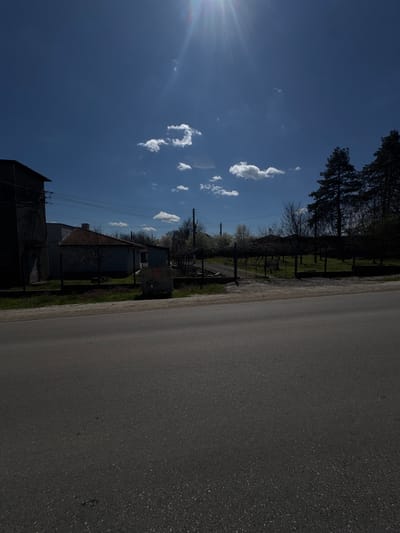 The image depicts an outdoor scene in Dobrotitsa, Bulgaria, featuring a rural road stretching to the right with a dark asphalt surface. A clear blue sky with scattered, small white clouds is visible. A row of houses and vegetation lines the left side of the road. Power lines run parallel to the road. Trees are visible in the background. The sun is positioned to the upper left, creating a bright glare. The scene appears to be taken on a sunny day.