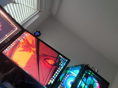 The image presents a low-angle view of a workspace, likely an indoor setting. Visible elements include two computer monitors displaying digital content and a computer case featuring illuminated fans. The screen on the left appears to show an anime-style image with red and orange hues. The computer case is lit with blue, green, and purple LEDs. Natural light is entering through a window at the top of the frame.
There are no people visible in the image.
The provided text does not include sufficient information to confirm the location as Havixbeck, Germany. Therefore, the description is based solely on the visible contents of the image.