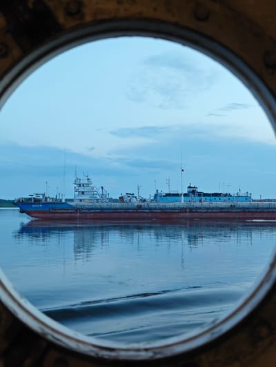 This image, taken through a porthole, shows a long, narrow barge with a red hull and a blue upper deck. The barge is on a calm body of water, likely a river, with its reflection clearly visible. In the distance, another, smaller vessel is moored alongside the barge. The sky is overcast with soft blue and white clouds, suggesting it is either early morning or late evening. The water is still, with only a slight ripple visible in the foreground, indicating a peaceful and serene atmosphere. The scene is set in Vorogovo, Russia, a location known for its waterways. There is text visible on the blue side of the barge, which appears to be "БН-14", likely a vessel identification number. The overall mood is one of quiet contemplation and the vastness of nature.