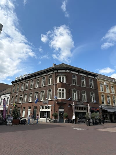 The image depicts a street scene in Roermond, Netherlands, with a focus on the facade of a brick building housing a restaurant named "RHODOS". The building has multiple floors with numerous windows, many of which are ornate. Flags, one with a blue and white pattern, are visible on the building. In front of the restaurant, tables and chairs are set up outdoors under umbrellas, suggesting a cafe or dining area. Adjacent to "RHODOS" are other businesses, including "Pâtisserie" and "de Kroon". Several people are walking on the brick-paved sidewalk in front of the buildings, and a scooter and bicycles are parked nearby. The sky above is blue with scattered clouds. The overall scene suggests a moderately busy urban environment on a pleasant day.