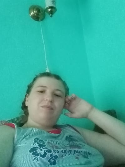 An indoor, eye-level, medium shot shows a woman with her head resting on her hand. The woman is looking towards the camera. She has medium brown hair pulled back and is wearing a sleeveless, striped shirt with a red neckline and a floral graphic. The background is a solid teal wall, with a ceiling light fixture visible in the upper left corner. There is no indication of the location being in Kitskany, Moldova.
