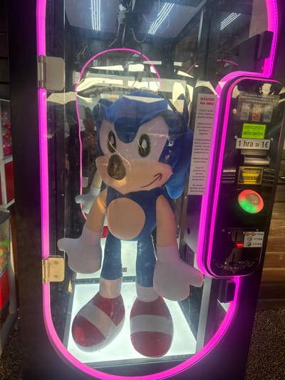 A close-up, eye-level shot captures a Sonic the Hedgehog plush toy inside a brightly lit arcade claw machine. The machine, adorned with pink neon lights, stands in what appears to be an indoor establishment, possibly a game arcade or a shopping mall, in Zamarovce, Slovakia. The plush toy, the main subject, is positioned centrally, its blue fur, iconic red and white shoes, and white gloves are clearly visible. The machine's interface is on the right, featuring a coin slot, buttons, and instructions in Slovak. A sign above the coin slot reads "NÁVOD NA HRU" (Instructions for Play). The machine is designed to allow players to attempt to win prizes, like the plush toy, by operating a mechanical claw. The background is a blurred reflection of the surroundings, suggesting a public space with people and other machines.