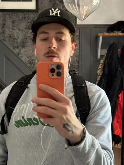 A young man takes a selfie in a mirror. He wears a grey sweatshirt with green lettering, a black baseball cap with a white "NY" logo, and has a black backpack strap across his chest. He is holding an orange cell phone. A tattoo is visible on his left hand. The background includes a framed picture on the wall, light grey wallpaper with a textured pattern, and a clothing rack with various garments. The location is not discernible as Hitchin, United Kingdom.