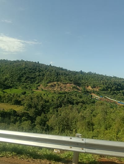 The image shows a landscape in Muthambi ward, Kenya. A green, densely forested hillside dominates the view, with an area of exposed soil or excavation visible on the slope. Lower down, there's more scattered vegetation and a glimpse of some structures or buildings. In the foreground is a metal guardrail, indicating the perspective is from a road. The sky is a clear blue with some clouds.