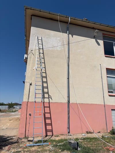 The image depicts an exterior view of a building located in Karageorgievo, Bulgaria. A tall, silver-colored ladder stands upright against the building's light beige and pink-painted facade. The ladder has orange straps and is positioned near a downspout. Various cables and wires are visible running along the building and on the ground. The ground around the building is unpaved and appears to be undergoing some form of construction or renovation, with debris, including bricks and white wiring, scattered on the ground. In the foreground, a black cylindrical object, possibly a bucket or pipe, lies on the grass. The sky above is clear and blue, indicating fair weather conditions. The building has visible windows and a gutter system.