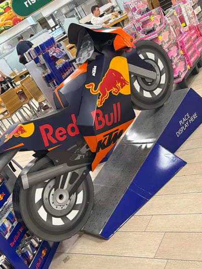 The image captures a detailed view of a Red Bull branded KTM motorcycle display, likely in a supermarket or retail store environment. The display is constructed from cardboard and features the iconic Red Bull logo and KTM branding. The motorcycle is positioned on a stepped platform. In the background, several people are visible, some appearing to be customers or staff. One person is seen at a counter, possibly preparing food. Other individuals are seated at tables in what appears to be a dining area. Stacks of Red Bull cans are visible on shelves to the left of the display, and boxes of toys, possibly dollhouses, are stacked on shelves to the right. The overall setting suggests a commercial space with a variety of products and services available. The flooring is a light-colored wood-look laminate. The context of Due Carrare, Italy, indicates this scene is located within that specific town.