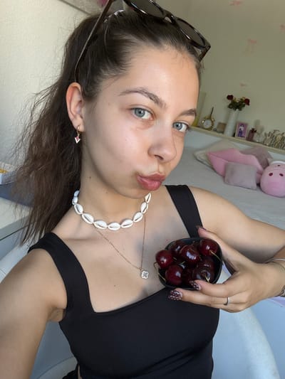 A young woman with fair skin and light blue eyes is holding a small bowl of dark red cherries, posing for a selfie indoors. She has brown hair, sunglasses perched on her head, and a seashell necklace. She wears a black top. Her nails are painted dark brown with white polka dots. A ring is visible on her finger. The background includes a shelf with decorative objects, a pink stuffed animal, and a pillow. The location cannot be determined as Bruchköbel, Germany, from this image.