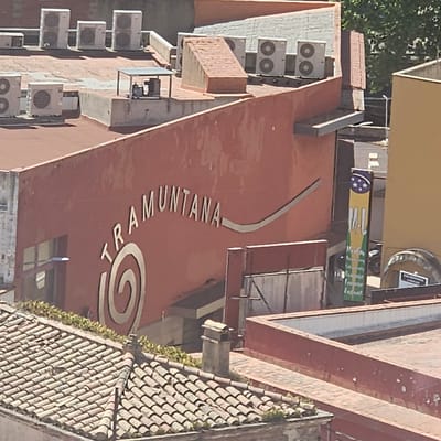This is an exterior shot of a building in La Jonquera, Spain. A salmon-colored wall displays the word "TRAMUNTANA" in silver lettering. Near the beginning of the word is a silver spiral design. Air conditioning units are visible on the building's facade. Above the word Tramuntana, there is a banner with what appear to be flags from Brazil, with some lettering in green and yellow. To the right is a brown-tiled roof with some plant life. The overall angle of the image appears to be from a slightly elevated position.