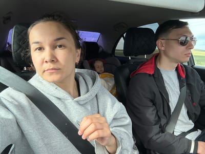 Inside a vehicle, likely in Sukhtelinskiy, Russia, three individuals are present. In the front passenger seat, an adult female with dark hair pulled back and brown eyes looks directly at the camera. She wears a light grey hooded sweatshirt and a black seatbelt, which shows minor fraying. A small gold hoop earring is visible on her left ear. In the driver's seat, an adult male with short dark hair is seen in profile, looking forward and to the right. He wears reflective aviator-style sunglasses, a grey t-shirt, and a black zip-up jacket with a visible red lining and a black-and-white striped cuff on the sleeve. He is also secured by a black seatbelt. In the rear passenger seat, an infant or young child is asleep in a car seat, wrapped in a yellow and white floral-patterned blanket, with dark, curly hair visible. The car's interior features dark upholstery, including quilted headrests, and the rear windows appear to have a purple tint or reflection. Through the vehicle's windows, a vast, flat, green-brown steppe landscape with a partially cloudy sky is visible, with power lines stretching across the distant horizon.