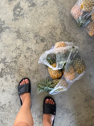 The image shows two pairs of black slide sandals on a concrete floor. A person's feet are visible, wearing the sandals. In the background, there are several pineapples in clear plastic bags. One bag is closer to the foreground and contains multiple pineapples, with their green leafy tops visible. Another bag of pineapples is further in the background, on the left side of the image. The concrete floor appears worn, with some patches of discoloration and cracking. The lighting suggests it is daytime, possibly outdoors given the texture of the concrete. The overall scene appears casual and domestic, possibly in a yard or walkway in Hội Tín, Vietnam. There is no visible text in the image.