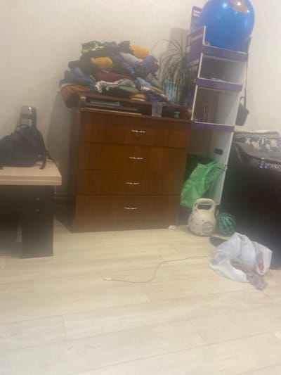 The image depicts an indoor scene, likely a room in a residential building. The focal point is a wooden dresser with three drawers, partially obscured by a large pile of clothing on top. To the left of the dresser, a small, dark-colored table holds a black backpack. A tall, white shelving unit stands to the right of the dresser, topped with a large blue inflatable globe. A potted plant with long, thin leaves is positioned behind the dresser and to the left of the shelving unit. On the floor in front of the shelving unit, there is a white kettlebell and a small, round, dark green object, possibly a decorative sphere or a small watermelon. A green plastic bag lies behind the kettlebell. Further to the right and closer to the foreground, a white plastic bag is open, containing some greyish material. A thin wire runs across the floor from the lower left to the mid-right of the image. The room has light-colored wood-plank flooring and pale walls. The lighting is dim, and the overall atmosphere is cluttered and somewhat disorganized. There is no indication of the city of Askarovo, Russia within the image itself.