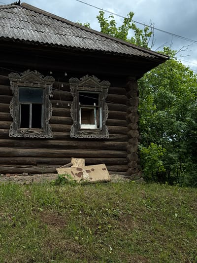 A low-angle, medium shot shows a partially visible, dark brown log cabin in Medvedka, Russia, with two ornate, wooden-framed windows. The top window has a reflection of the sky, while the bottom window appears dark. To the right of the cabin is a patch of green grass and foliage. A section of corrugated metal roofing is visible at the top left of the frame. Power lines run across the top left of the image. The sky is overcast. A piece of tan-colored debris rests against the side of the cabin.