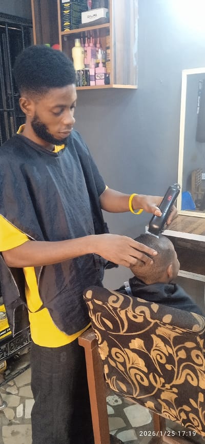 A barber with a beard, wearing a black and yellow cape and a yellow wristband, is giving a haircut to a customer. The barber is using an electric clipper to cut the customer's hair. Various hair care products are on a shelf in the background. A chair with a patterned seat is visible. A generator is also visible in the background. The date "2026/1/25" is printed on the bottom right corner.