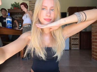 This image depicts three young individuals within an indoor residential environment located in Quilmes Oeste, Argentina, illuminated by natural light during daytime.
In the foreground, a female with long blonde hair, blue eyes, and a septum piercing is positioned in a low stance, possibly kneeling or squatting. She is wearing a black strapless top. Her right arm is extended, holding a device for a selfie, while her left hand is raised, palm flat against the side of her face. Her right forearm displays a small smiley face tattoo and multiple bangles, including one ornate silver-colored piece.
In the mid-ground, behind the primary subject, two other individuals are present. To the left, a male with dark hair and glasses wears a black t-shirt with white graphics. He stands behind a wooden table on which a 2.25-liter plastic bottle of "SODA" with a blue label is placed. To his right, a female with reddish-brown hair styled in two buns wears a white tank top, camouflage-patterned trousers, and a studded belt. She has visible tattoos on her left arm, and her left hand is positioned near her mouth. Both individuals appear to be interacting, though their specific actions are not fully clear.
The environment features light brown tiled flooring. A dark wooden cabinet, displaying glassware and topped with artificial green plants and red flowers, is visible behind the male. To the right, a brick wall and dark wooden kitchen cabinets are present. An air conditioning unit is mounted on the wall above the wooden cabinet.