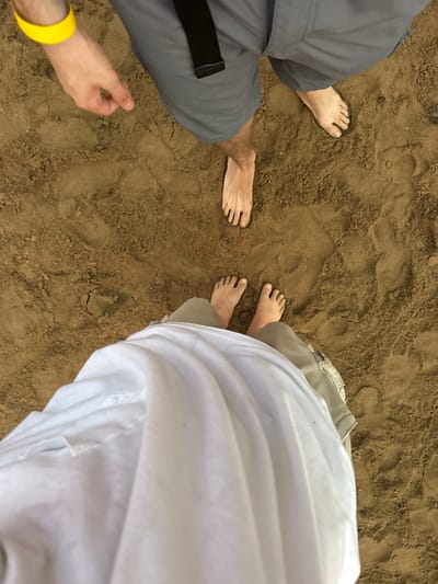 The scene depicts two individuals standing barefoot on reddish-brown sand, characteristic of an outdoor environment such as a beach or sandy riverbank, located in Ban Mae Ping, Thailand.
In the foreground, the lower body of one person is visible, wearing light-colored (possibly khaki) shorts, largely covered by a flowing white garment. Their bare feet are placed together on the sand, showing distinct toe impressions.
In the background, positioned further away, is the lower body of a second person. This individual wears gray shorts secured by a black belt. Their bare feet are spread apart, with the right foot slightly forward. A left arm, bare from the elbow down, extends from the top left corner, adorned with a bright yellow wristband. The hand is partially clenched.
The sand beneath both individuals is disturbed, showing multiple footprints and indentations from prior movement. No other objects or specific actions beyond standing are discernible.