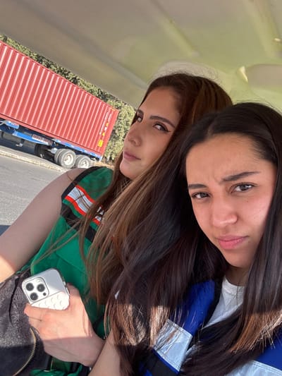 The image captures two adult females seated inside a light-colored vehicle, likely an open-sided utility vehicle, under bright daylight conditions. The woman positioned to the left and slightly behind has long, brown hair with lighter highlights, wears a green garment with white and orange/red reflective detailing, and holds a light-colored smartphone with a prominent camera array and textured case in her right hand. Her gaze is directed towards the left. The woman closer to the foreground on the right has long, dark brown hair, wears a blue and white top over a white t-shirt, and looks directly at the camera with a neutral to slightly serious expression. In the background, a large red shipping container is visible on a blue chassis with multiple wheels, partially displaying the text "MAVZ 20125". The container rests alongside a paved road, backed by green foliage and trees. The scene is located in Sanctorum, Mexico.