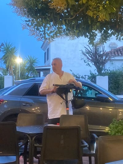 An outdoor scene at dusk, likely in San Pedro De Alcántara, Spain, shows a middle-aged man standing with his hand on the handlebars of a black electric scooter. He is wearing a loose-fitting white shirt with a lace-up neckline and white pants. The man has short grey hair and is looking towards the right of the frame. Behind him, a dark grey SUV is parked. In the foreground, there are dark chairs and tables, suggesting a cafe or restaurant setting. Palm trees and foliage are visible in the background, along with a white building with a red-tiled roof and windows. The sky is a deep blue, indicating twilight. The lighting suggests artificial lights are also present, as seen by reflections on the car and the illuminated street lamp in the upper left.