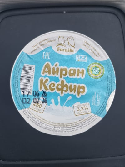 This is a close-up, top-down shot of a container of Ayran Kefir, a dairy product, likely taken in a kitchen or on a table. The container is round with a light blue lid featuring a textured pattern. The lid prominently displays the brand "Familk" in stylized gold lettering, with the product name "Айран Кефир" (Ayran Kefir) also in gold.

Key details on the lid include:
- A barcode on the left side.
- A stamp indicating "EAC" and "KZ" (Kazakhstan).
- A circular "HALAL" certification logo.
- Nutritional information in both Kazakh and Russian, indicating 3.2% fat content.
- The volume "250 г" (250 grams) and the word "КӨЛЕМІ" (Volume) in Kazakh.
- A date stamp showing "17.06.26" and "02.07.26", likely indicating manufacturing and expiry dates.
- Text in Kazakh and Russian provides details about the product's safety and management systems, its origin from the "Zhanaozen Dairy Plant" in Kazakhstan, and its nutritional values. The text also mentions storage instructions at 4±2°C.

The background is dark and out of focus, suggesting the container is the sole subject of the image. The lighting appears to be artificial, highlighting the product's packaging.