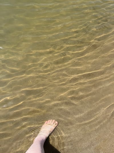 A human foot, partially submerged in shallow, clear water, is visible in the lower left quadrant of the image. The foot is fair-skinned and shows toes. The water creates rippling light patterns on the sandy bottom. The environment appears to be a body of water with a sandy shore, possibly a lake or river. The location is the city of Indiya, Moldova.