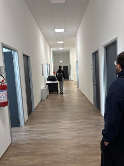 The main image, originally rotated, depicts a bright, modern indoor hallway, likely within an institutional or office building in Camerino, Italy. The perspective is from one end, looking down its length.

The hallway features light wood-grain laminate flooring laid in a linear pattern, clean white walls, and a white suspended ceiling fitted with numerous rectangular recessed light panels that provide ample illumination. Dark grey doors with silver frames line both sides of the hallway, most of which are closed.

In the immediate foreground, the upper body of a person wearing a dark blue top and glasses is partially visible on the right. Further down the hallway, another individual is seen from behind, walking away from the viewer. This person wears a black and white jacket, light grey trousers, and a black backpack.

Near the person in the middle distance, a large black printer or copier machine sits on the floor next to a white rectangular air conditioning or ventilation unit. On the left wall further down, there are white tiled panels or sections. A green exit sign is visible further along the hallway.

In the extreme foreground, a red fire extinguisher is prominently mounted on a white surface. Visible text on its label includes "FIRE EXTINGUISHER", "INSTRUCTIONS FOR USE", and partial regulatory information such as "PN EN 3-7+A1".

The scene suggests a functional, well-maintained, and brightly lit interior space.