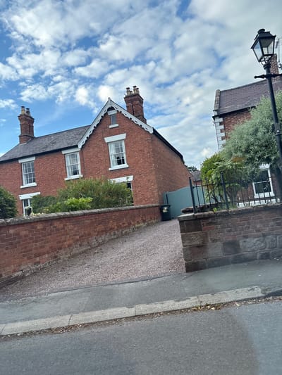 The image shows a red brick house with white-trimmed windows and chimneys. A gravel driveway leads to the house, bordered by a brick wall. There is a lamppost on the right side of the image, and a pale blue gate and a green bin are visible behind the wall. The sky is cloudy. The scene is likely a residential street in Chester, United Kingdom.