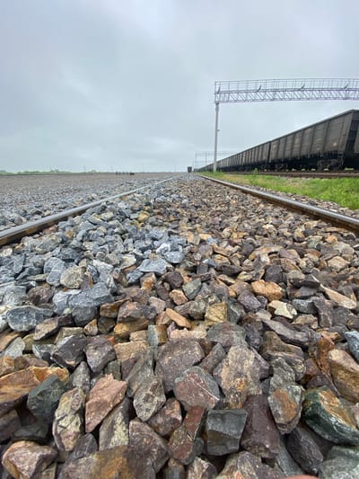 A low-angle, ground-level shot shows a railroad track with ballast in the foreground, extending into the distance under a cloudy, overcast sky. The ballast consists of various sized, angular stones in shades of gray, brown, and reddish-brown. To the right of the track, a long line of dark-colored, empty freight train cars is visible, stretching far into the background. Above the train cars, a metal overhead structure, likely for electrical wires, is present. In the far distance, a few utility poles and trees can be seen along the horizon. The environment appears to be an open, possibly rural or industrial area on the outskirts of Mayachnoye, Russia. There are no people visible in the frame.